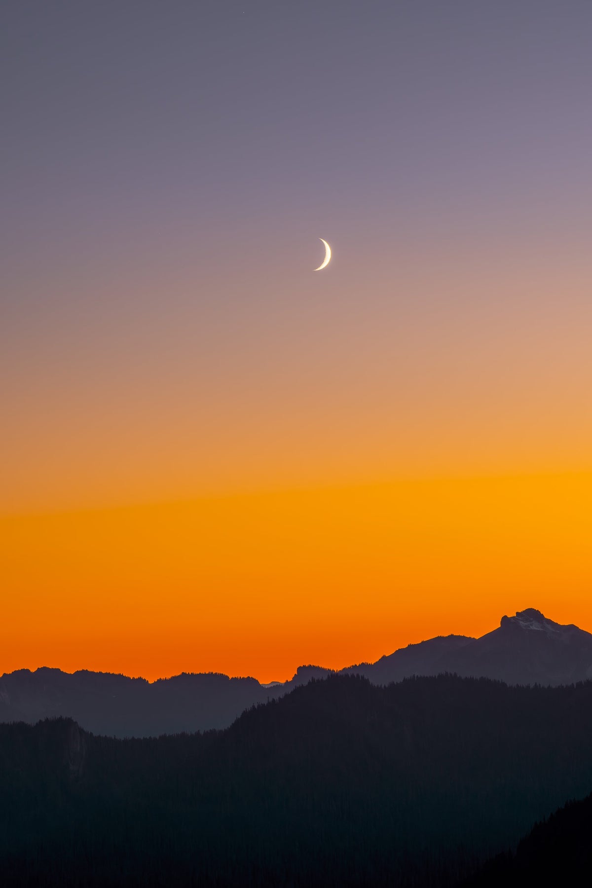 A thin crescent moon above an orange horizon over the Cascade Mountains