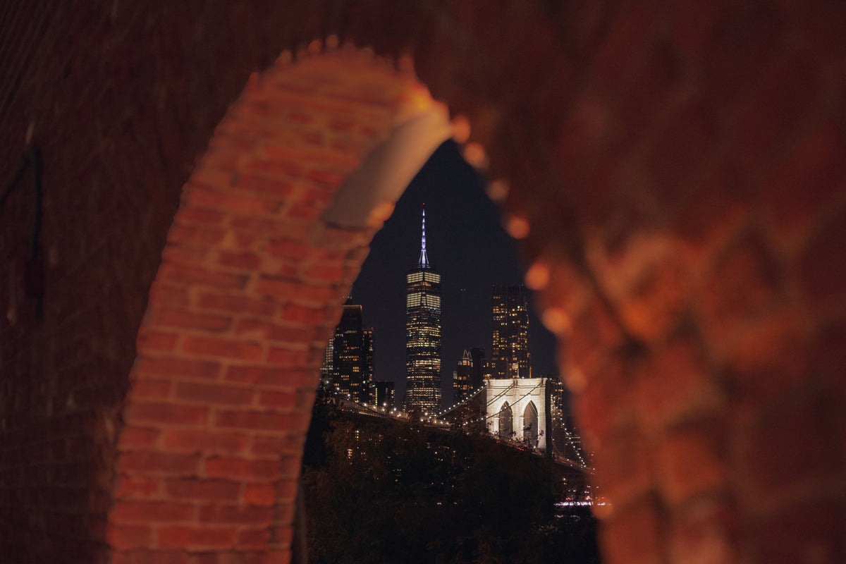 A brick arch in Brooklyn framing One World Trade Center and the Brooklyn Bridge at night