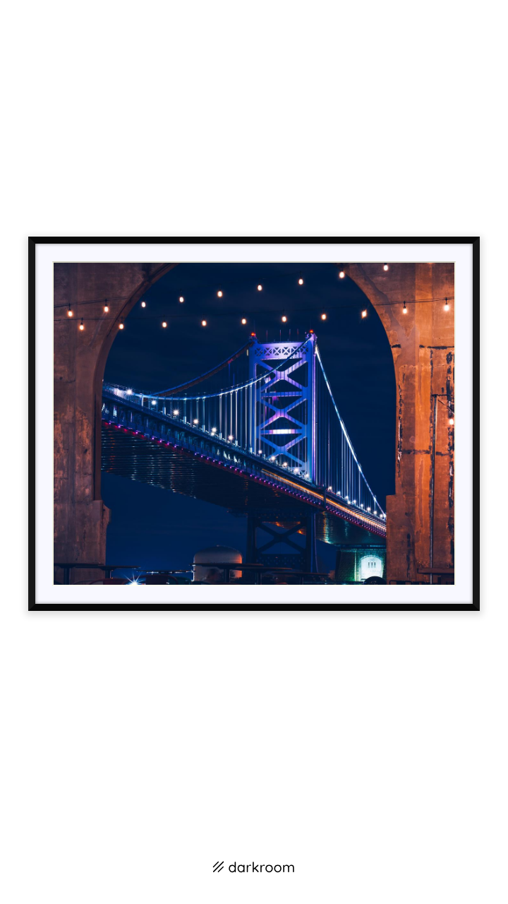 Black-framed print: Ben Franklin Bridge lit blue at night, framed by a brick arch and string lights
