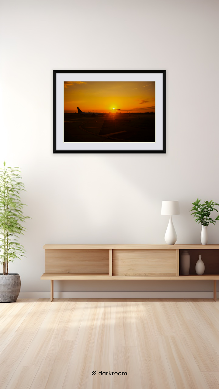 Black-framed print shown on a wall: A plane in silhouette on the runway as the sun sets